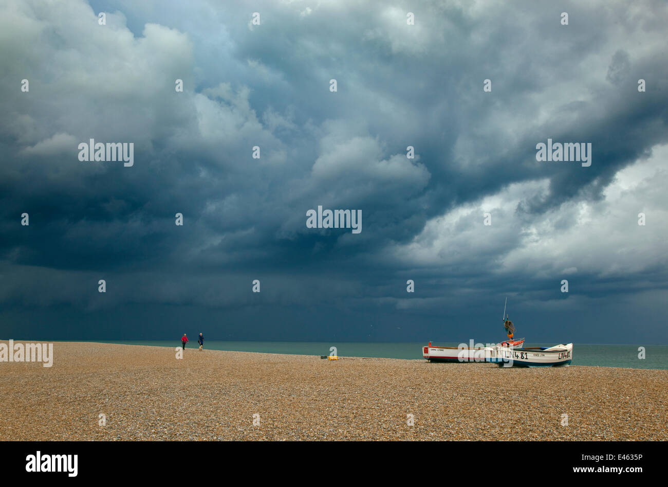 Due persone che camminano sulla spiaggia Cley vicino barche guardando verso Blakeney Point con nuvole temporalesche overhead, Norfolk, Regno Unito, Luglio 2011 Foto Stock