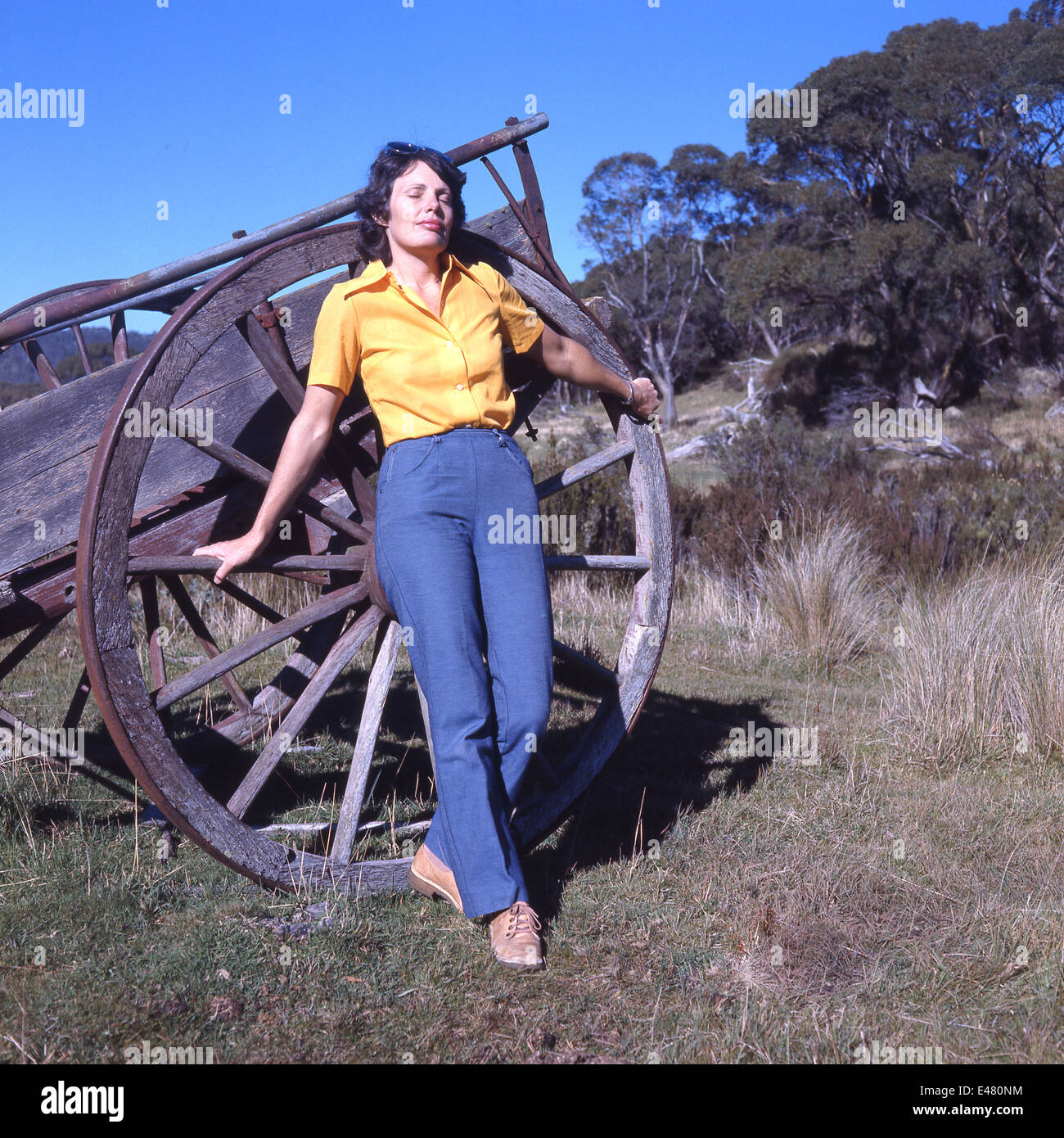 La donna in giallo camicetta e jeans blu appoggiata contro la vecchia fattoria ruota carrello godendo il calore del sole con gli occhi chiusi Foto Stock