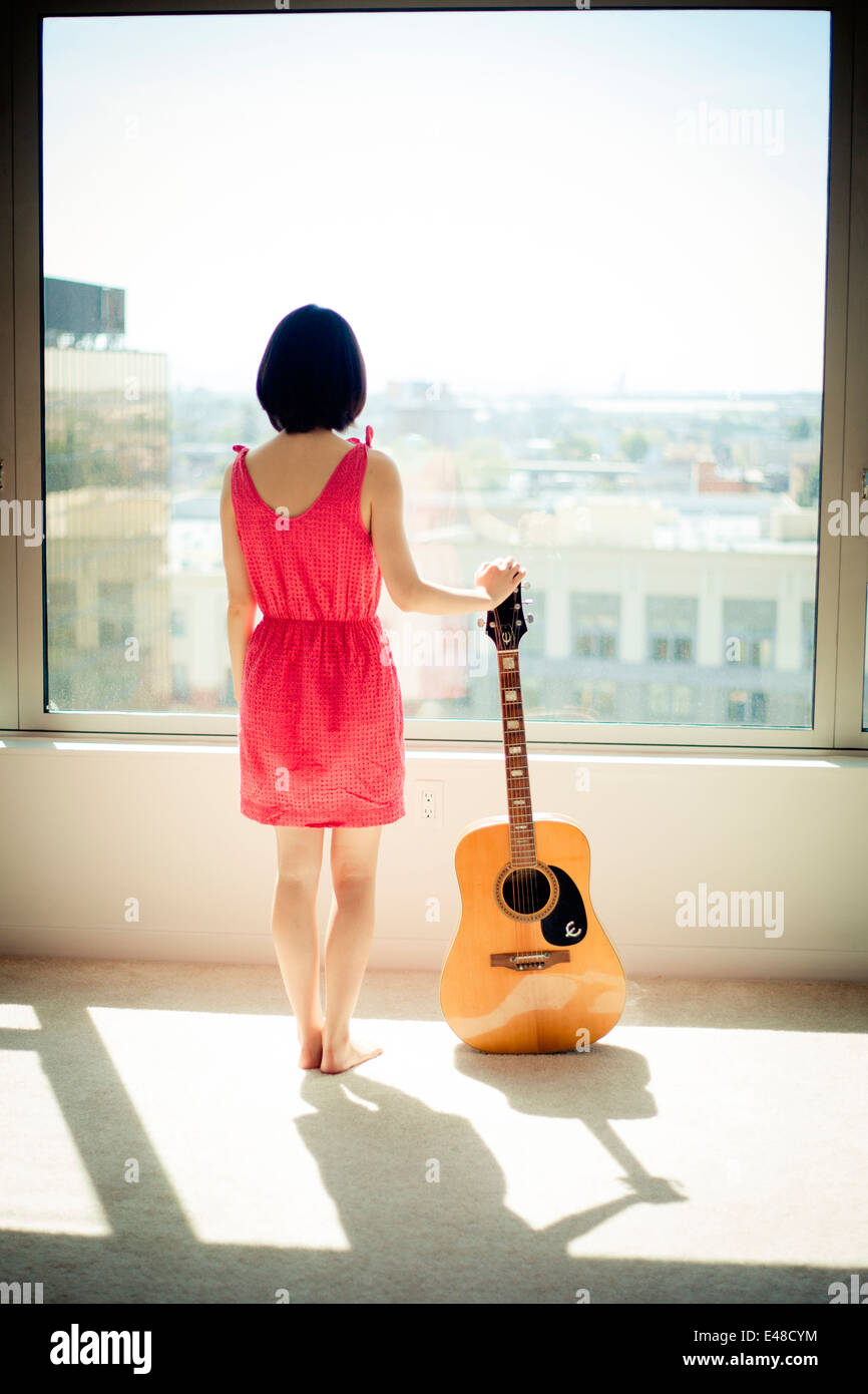 Vista posteriore della giovane donna in piedi con la chitarra e guardando attraverso la finestra Foto Stock