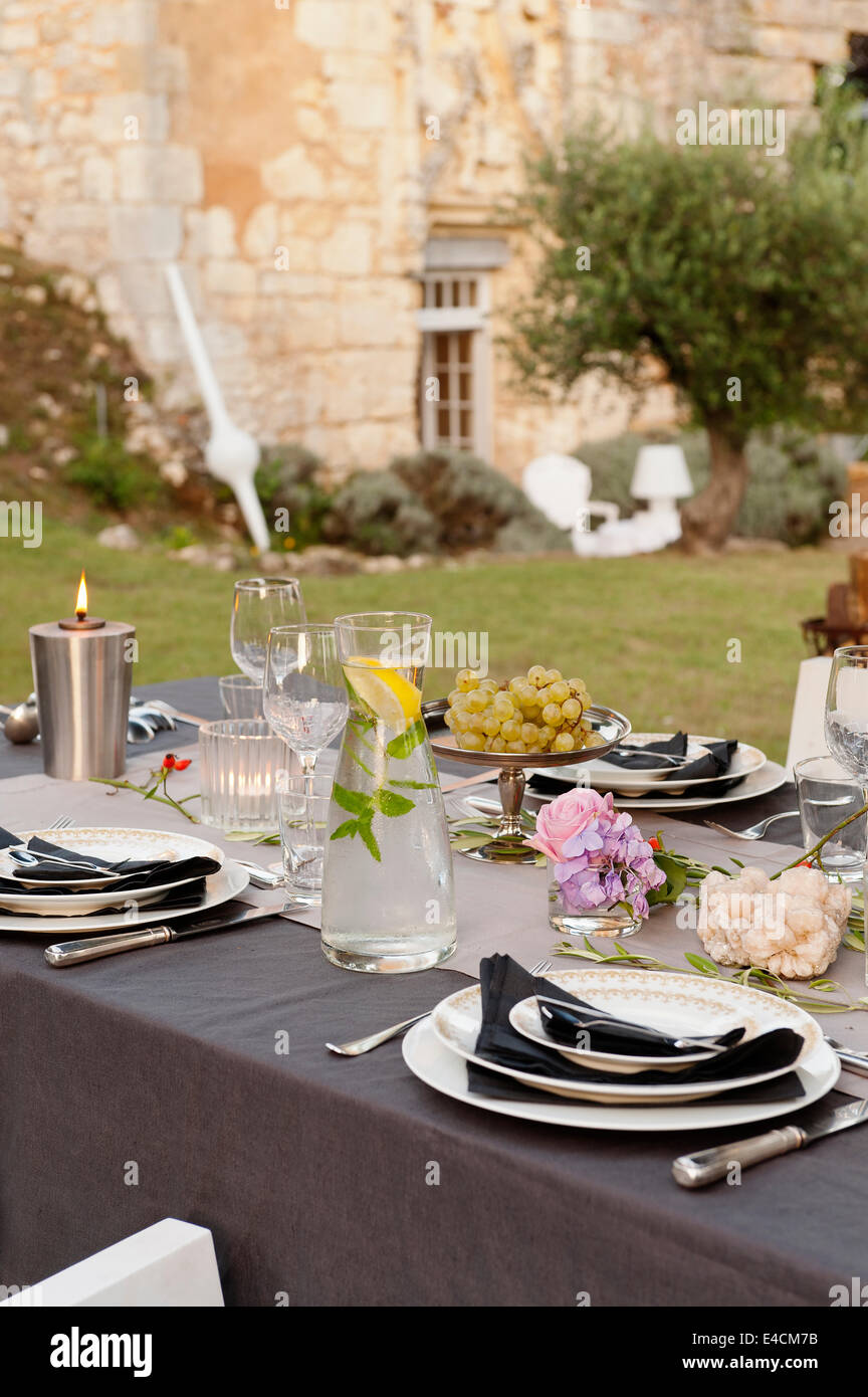 Tavolo esterno impostato per la cena con talee di rose e la brocca dell'acqua. Un albero di olivo e cappella del XII secolo può essere visto in backgrou Foto Stock