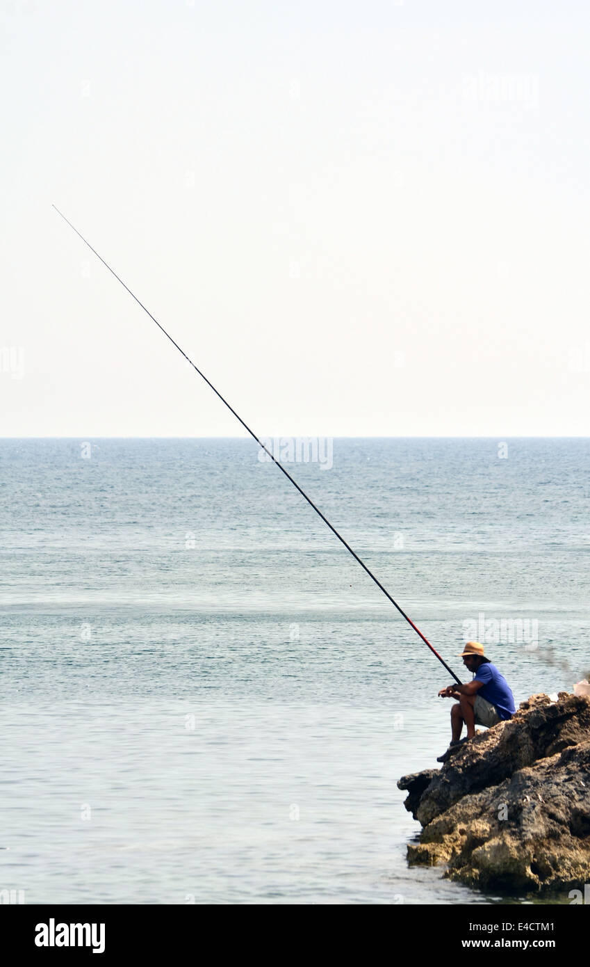 Pescatore solitario con una lunga asta sulle rocce al Coral Bay, Cipro Foto Stock