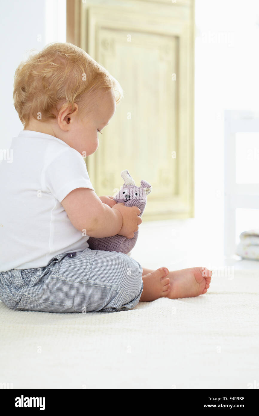 Baby boy (10 mesi) seduto sul pavimento afferrando a maglia giocattolo morbido Foto Stock