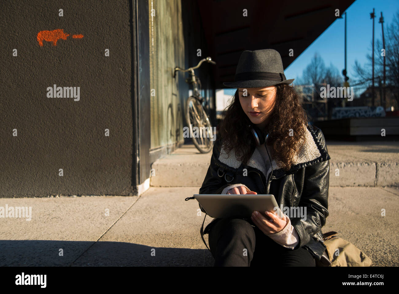 Ragazza seduta sul cordolo di marciapiede esterno, indossando fedora e utilizzando computer tablet, Germania Foto Stock