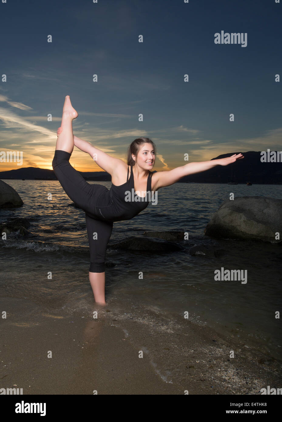 Un attraente giovane donna in nero spandex colpisce un yoga posa in onde di una spiaggia al tramonto. Foto Stock