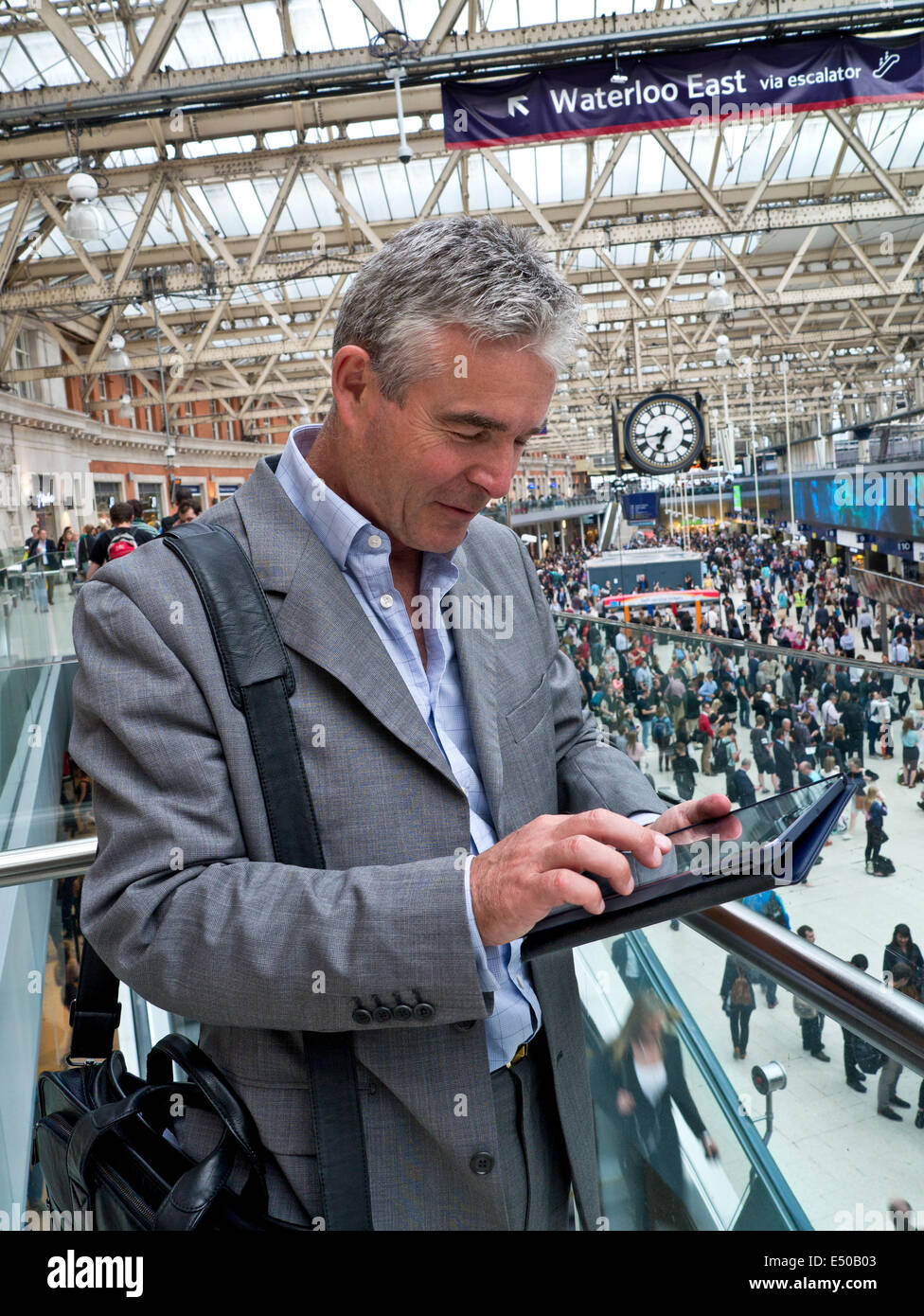 IPad Stazione ferroviaria uomo d'affari maturo alla stazione ferroviaria di Londra Waterloo usando il suo computer tablet iPad Air 'in movimento' con pendolari dietro Foto Stock