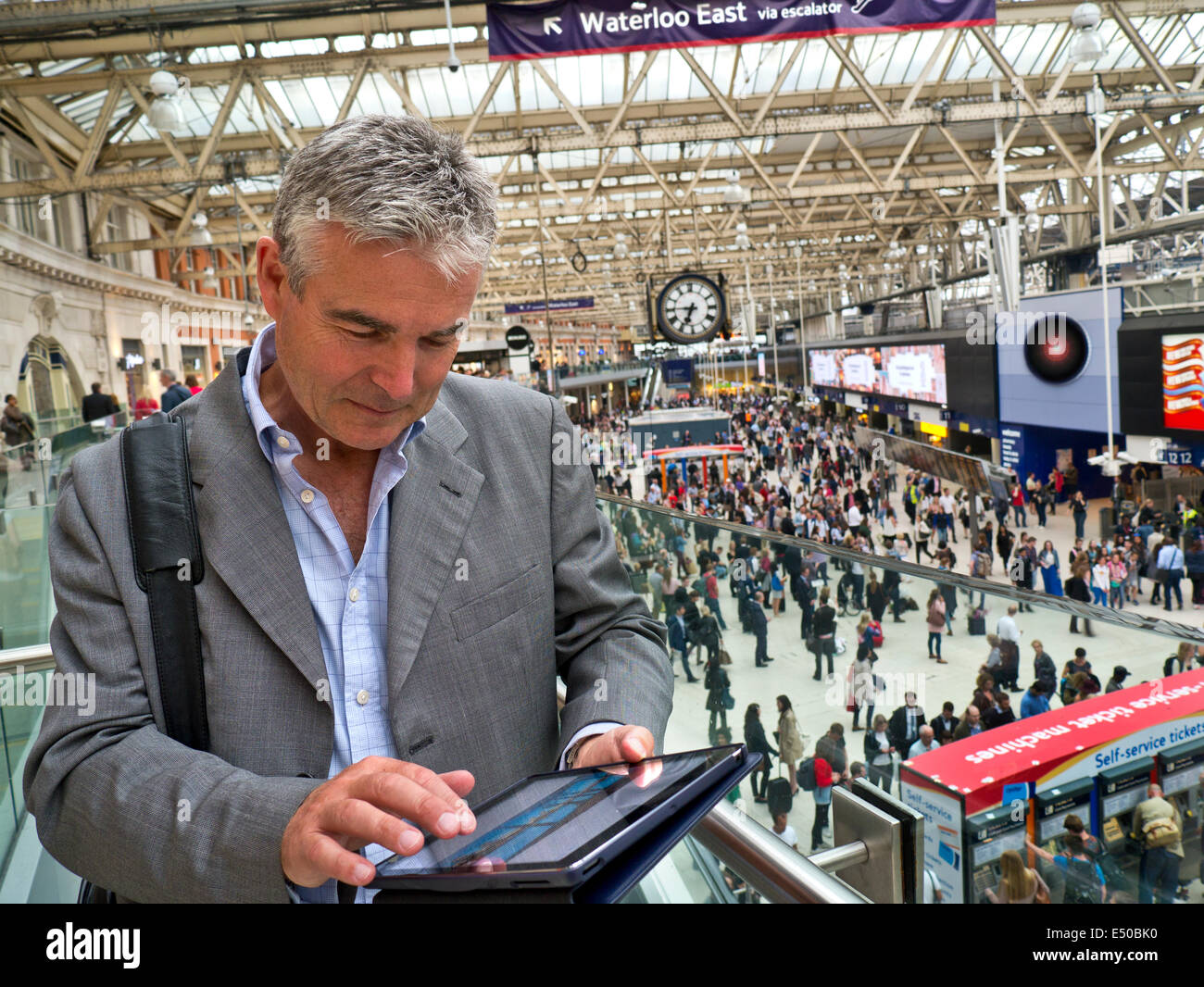 Imprenditore maturo su London Waterloo railway concourse usando il suo iPad aria compressa computer 'in movimento' con pendolari dietro Foto Stock