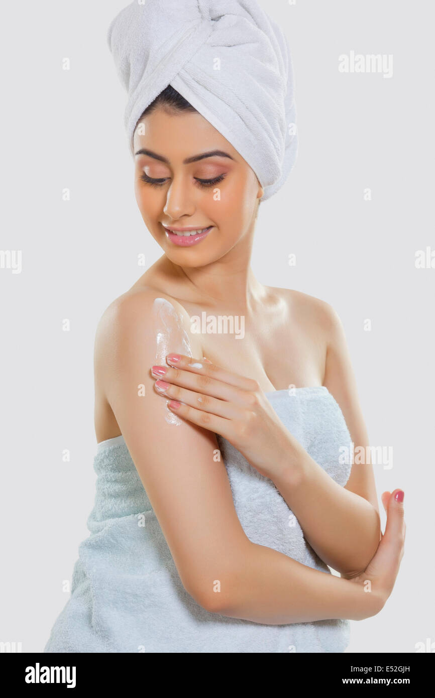 Bella donna avvolta in asciugamano di applicare una crema idratante su braccio su sfondo grigio Foto Stock