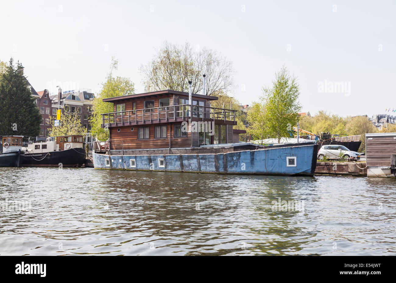 Casa insolita ormeggiata sulla riva di un canale di Amsterdam: casa in legno con una base in muratura, all'interno dello scafo di una barca blu Foto Stock