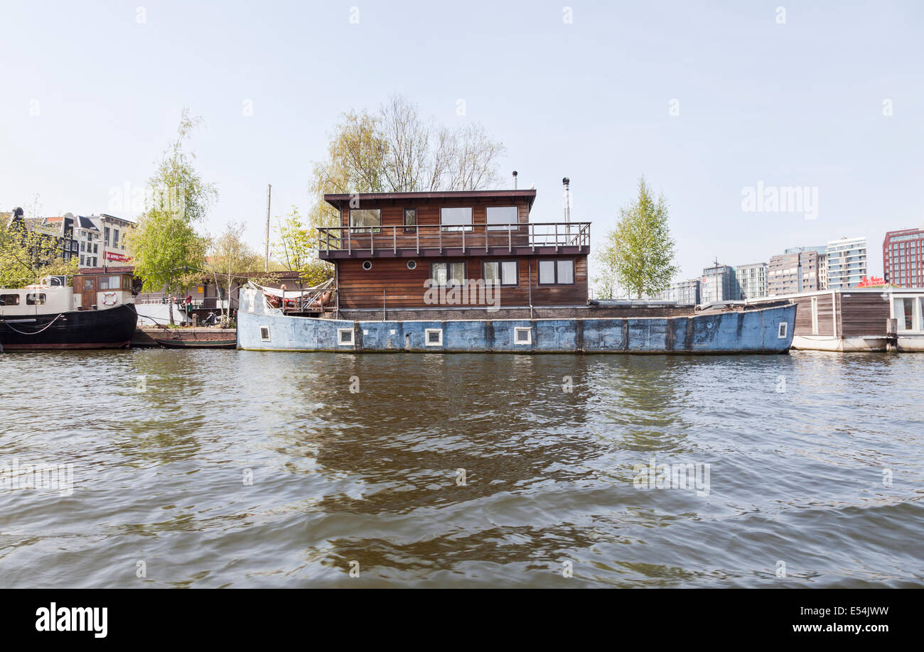 Casa insolita ormeggiata sulla riva di un canale di Amsterdam: casa in legno con una base in muratura, all'interno dello scafo di una barca blu Foto Stock