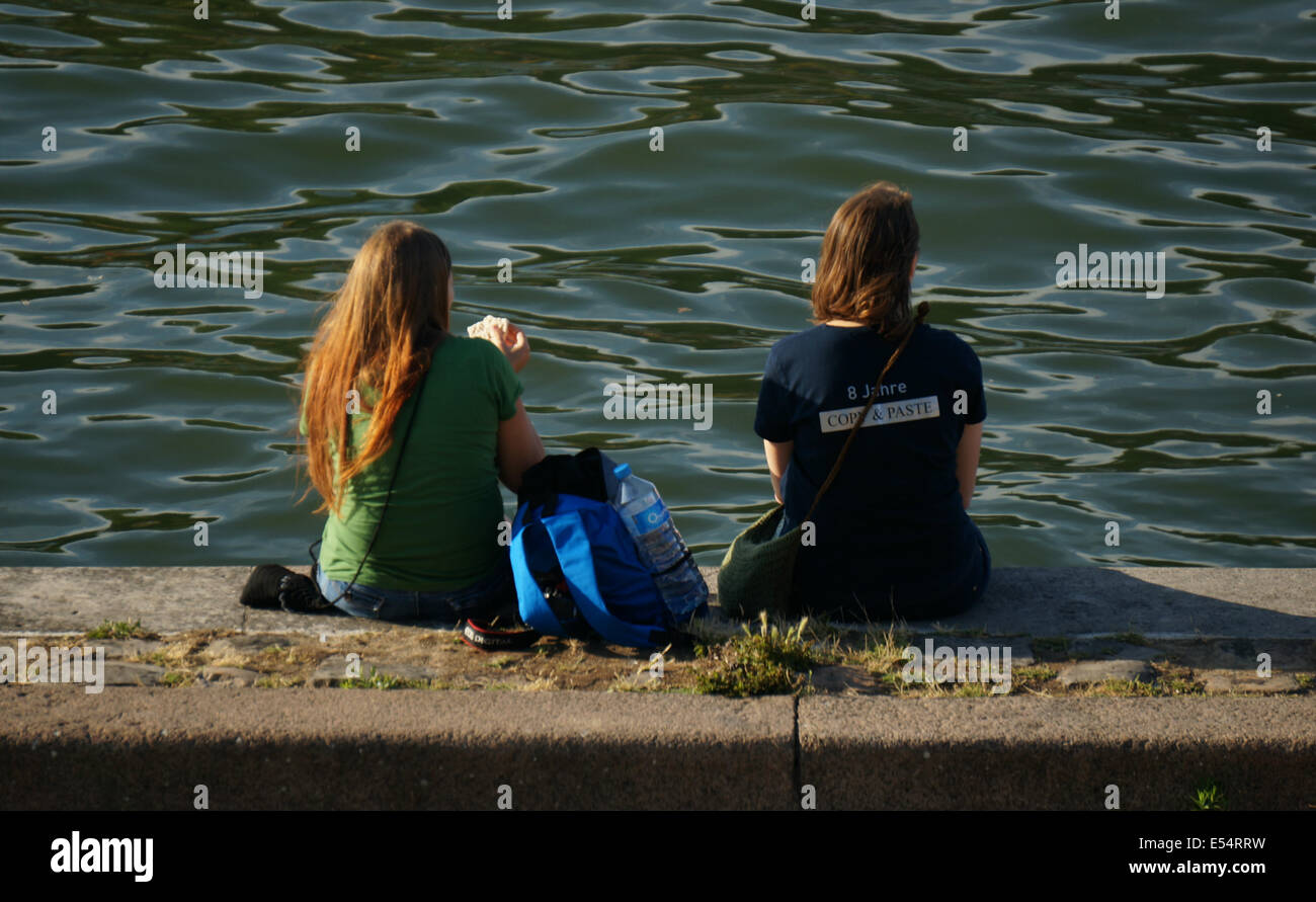 Vista posteriore di due donne turisti sedersi e rilassarsi con la chiara verdastra Seine Parigi; uno avente un panino come la sua cena Foto Stock