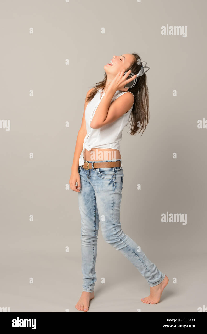 Ridere ragazza adolescente cantare godendo la musica dalle cuffie a lunghezza piena su grigio Foto Stock