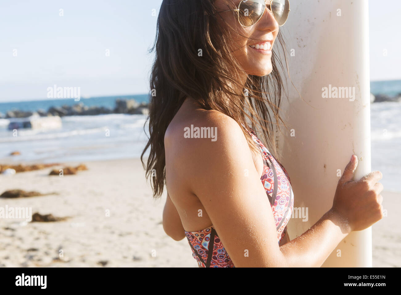 Giovani femmine surfer con occhiali da sole sorridente tenendo la tavola da surf in spiaggia Foto Stock