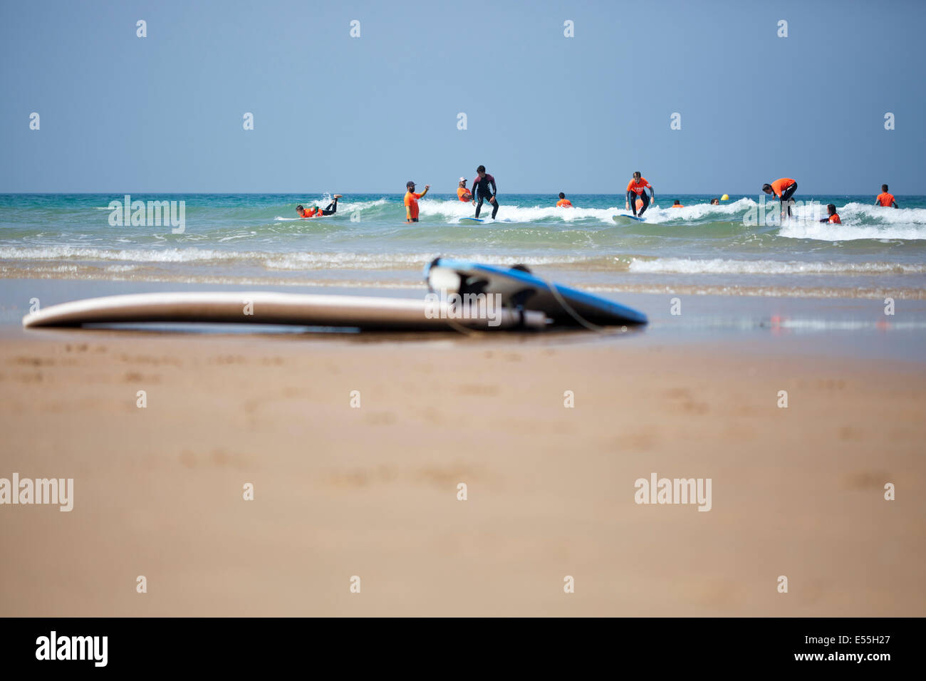 Gruppo di persone aventi lezioni di surf tra le onde su una spiaggia di sabbia in Spagna e tavole da surf in primo piano cielo blu in background Foto Stock