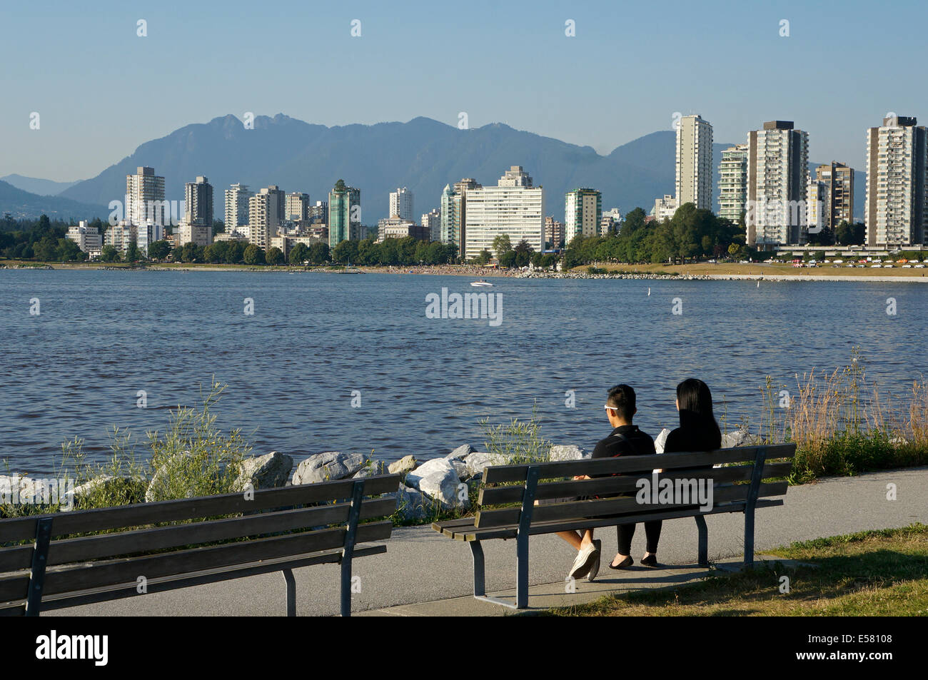 Giovani asiatici giovane su un banco affacciato sulla Baia di inglese con West End skyline e North Shore Mountains sullo sfondo, Vancouver, Foto Stock