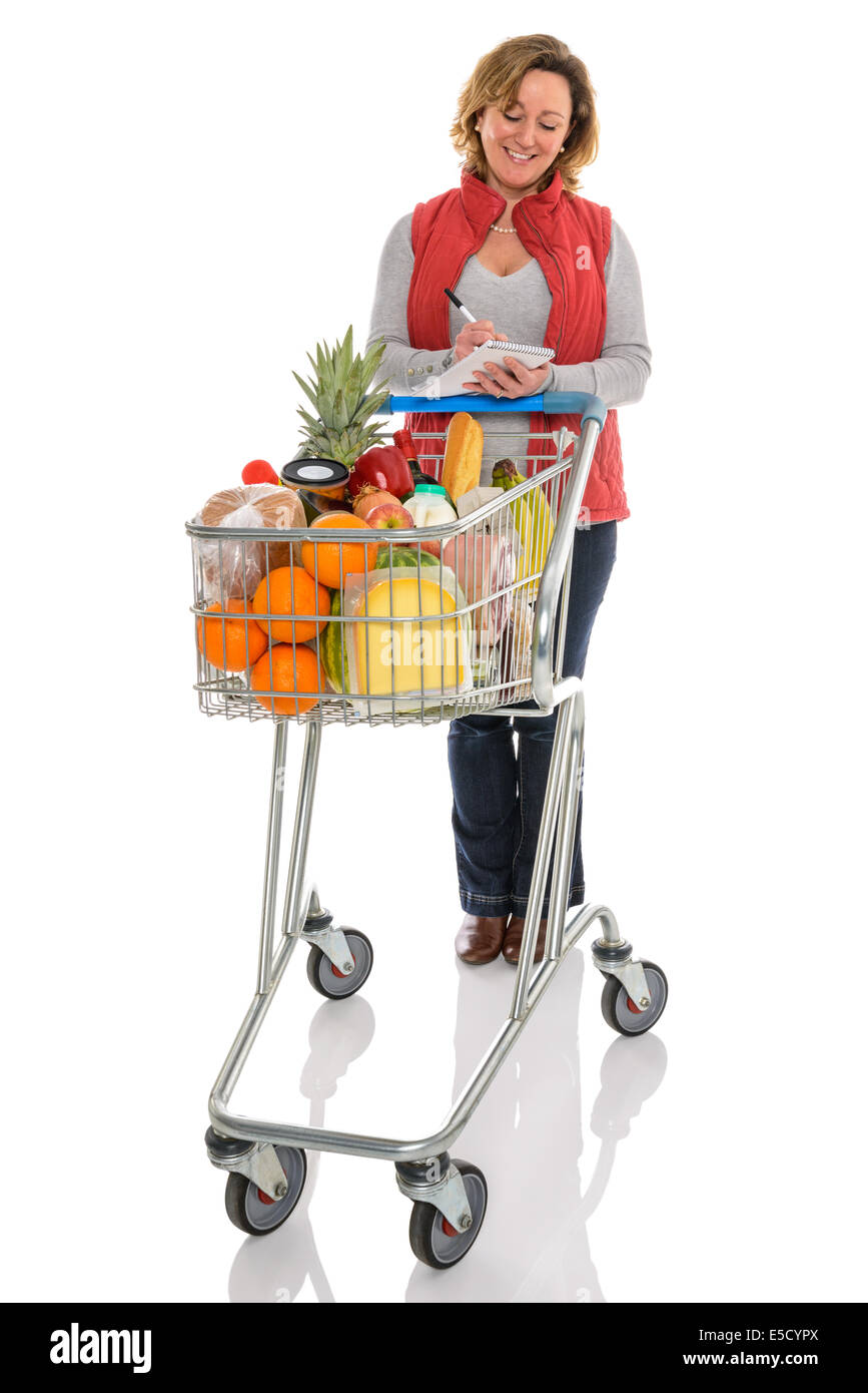 Una donna con un carrello per supermercati piena di cibo fresco controllando il suo shopping list, isolata su uno sfondo bianco. Foto Stock