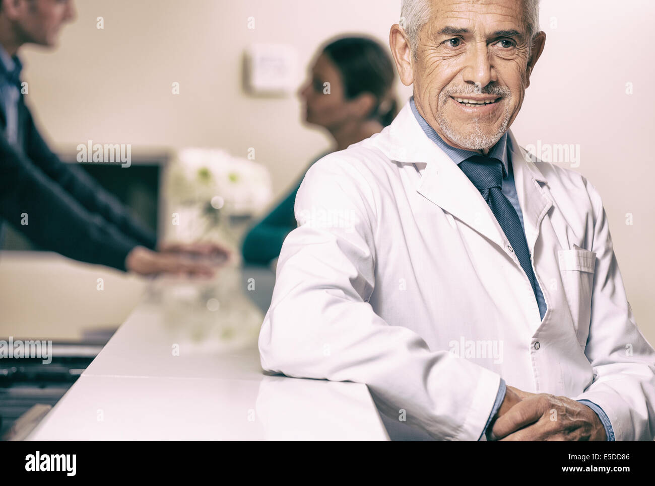 Sorridente maschio anziano medico sorridente all'ospedale reception, paziente di sesso femminile che parla con un giovane medico su sfondo. Foto Stock
