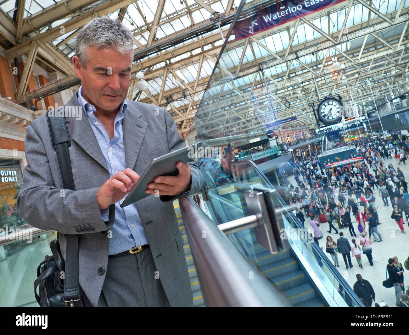 Imprenditore maturo su London Waterloo railway concourse usando il suo iPad aria compressa computer 'in movimento' con pendolari dietro Foto Stock