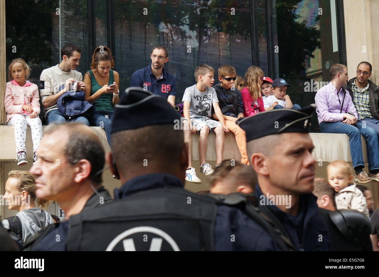 Famiglie e bambini seduti su un muro guarda la parata militare sul giorno della Bastiglia di Parigi, con 2 poliziotti in primo piano Foto Stock