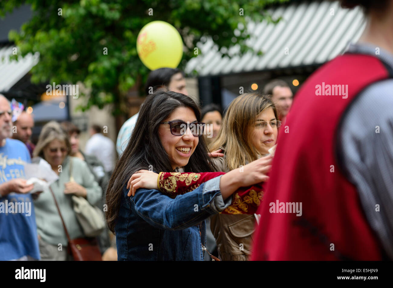 Giovane donna attraente in occhiali da sole si unisce con un greco ballo folk troupe, Marylebone street Fayre fiere estive, Londra, Regno Unito. Foto Stock