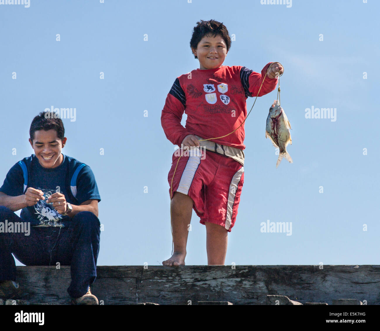 La visualizzazione delle catture. Un ragazzo visualizza un solo pesce pescato mentre un altro ragazzo ride. Entrambi sono sul bordo di un ponte di legno Foto Stock