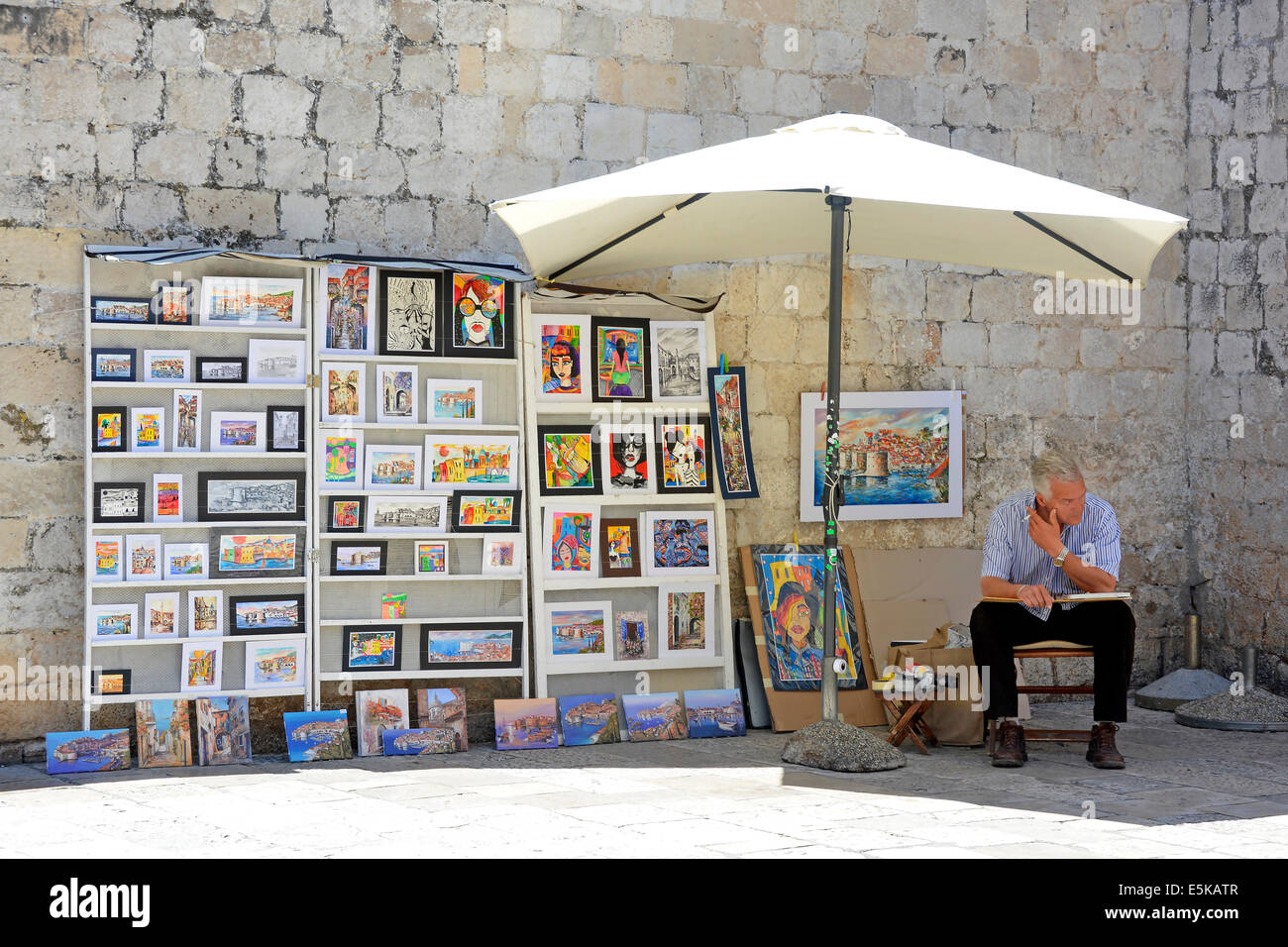 Uomo seduto in ombra sotto ombrellone dalla parete visualizzazione di illustrazioni per la vendita in area turistica in una calda giornata estiva Dubrovnik Croazia Dalmazia Adriatico Europa Foto Stock