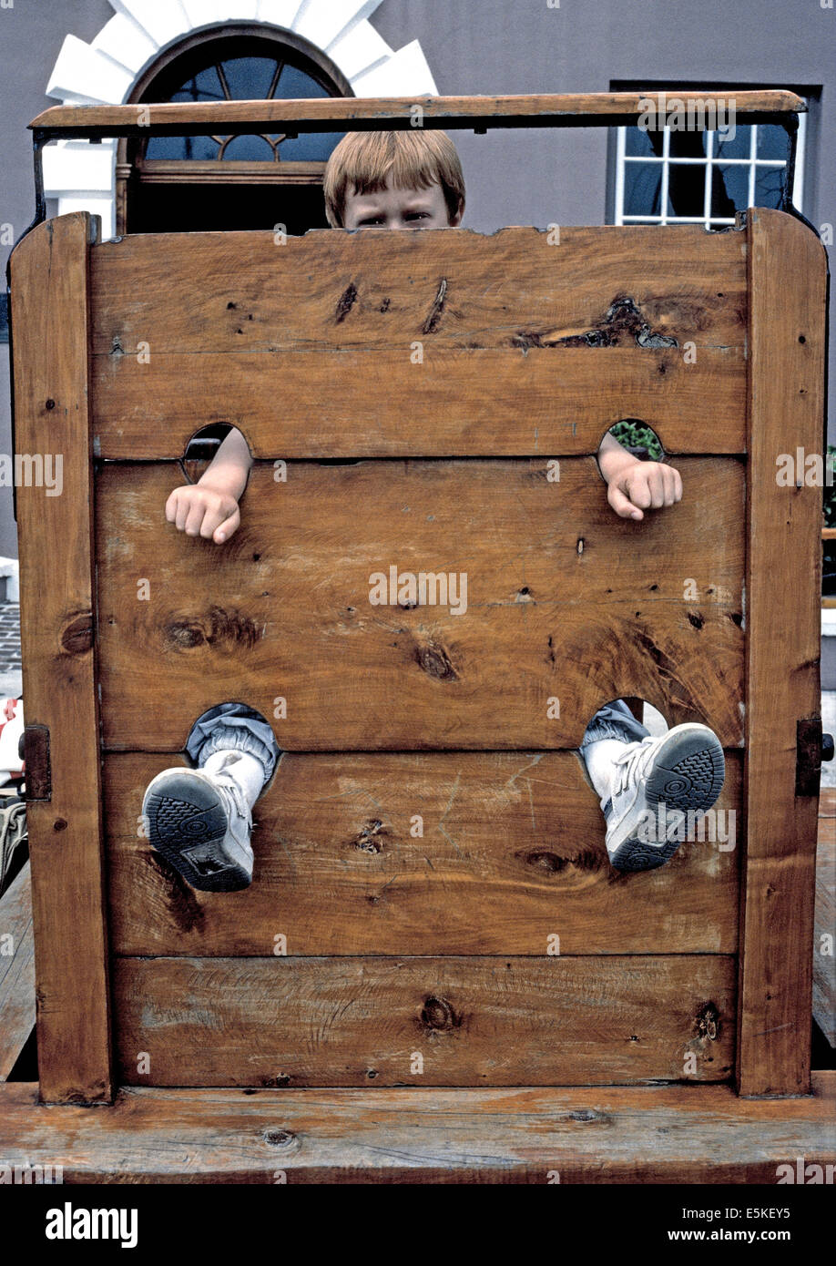 Un giovane ragazzo in posa per una foto con le mani e i piedi e sporgenti da una gogna, un comune pubblica punizione per crimini banali in early Bermuda. Foto Stock
