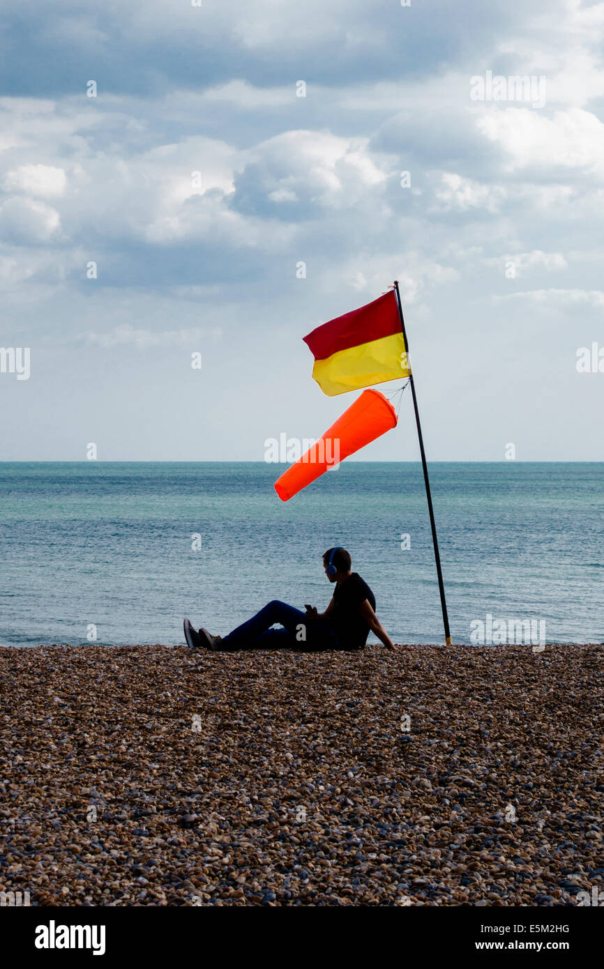 Un giovane ascolta la musica sulla spiaggia di ciottoli a Brighton, East Sussex, sotto bandiere che avvertono di pericolose condizioni di nuoto (UK) Foto Stock