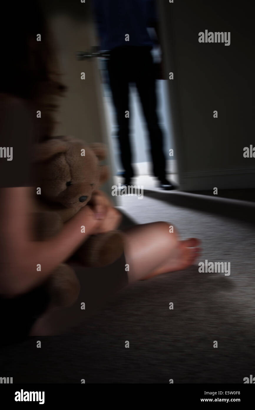 Anonimo uomo di entrare in una stanza buia, una giovane ragazza seduta in primo piano guardando verso l'uomo tenendo un teddy-bear. Foto Stock