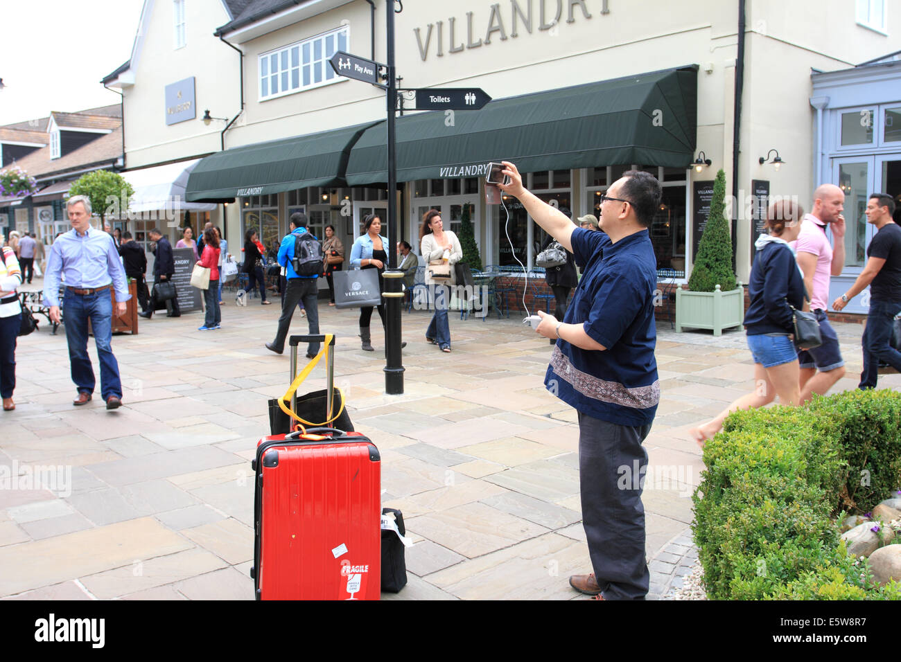 Cina uomo prendendo foto a Bicester Village,nome di marca negozi,turisti cinesi,prendendo foto,i bagagli, Oxford, Regno Unito Foto Stock