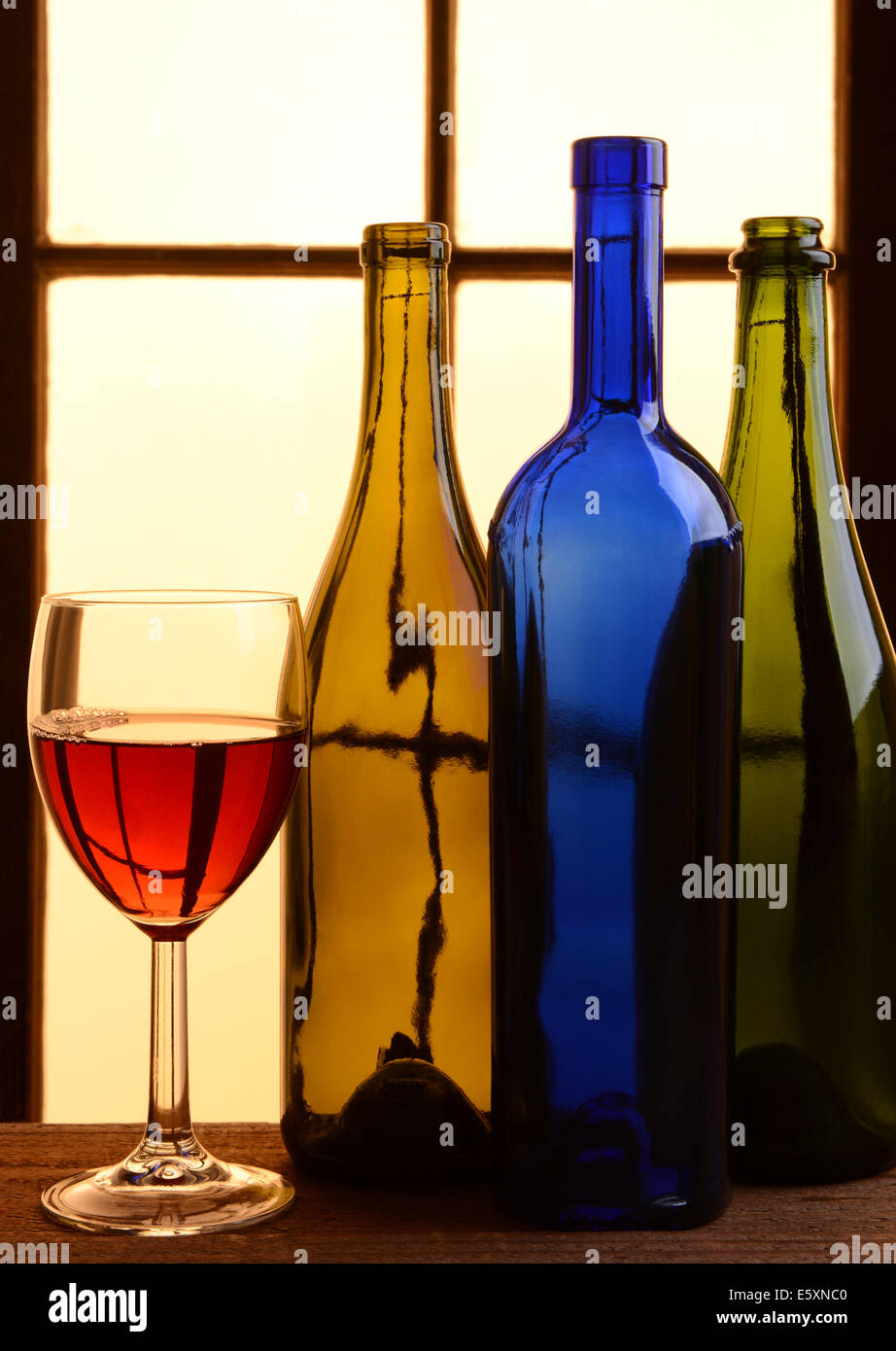 Un vino ancora in vita con toni caldi. Tre diverse bottiglie di vino e un bicchiere di vino rosso davanti a una finestra con una calda luce del sole. Foto Stock