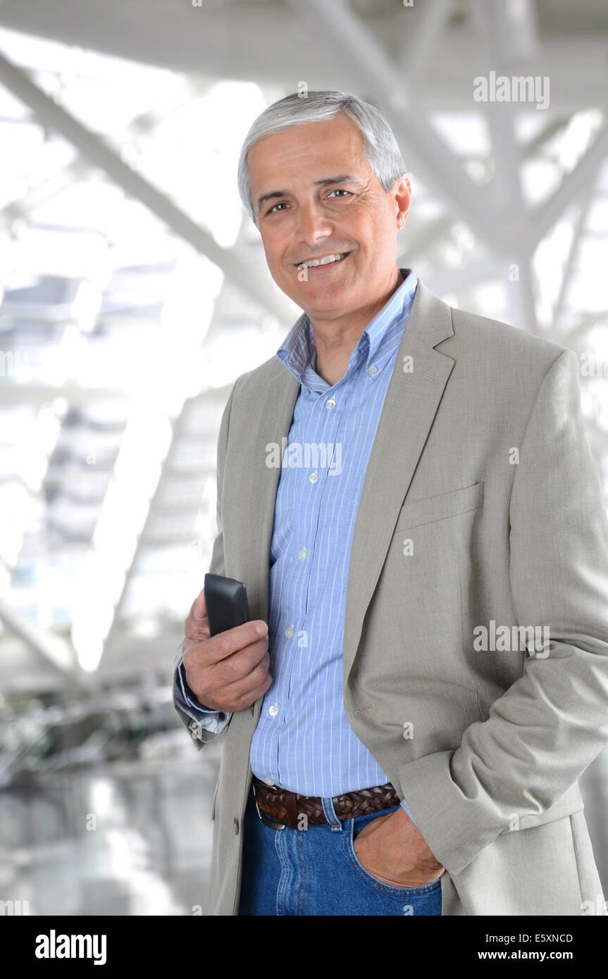 Un imprenditore informale con un telefono cellulare in piedi in un ufficio moderno edificio. L'uomo indossa jeans e un rivestimento di sport, una mano Foto Stock