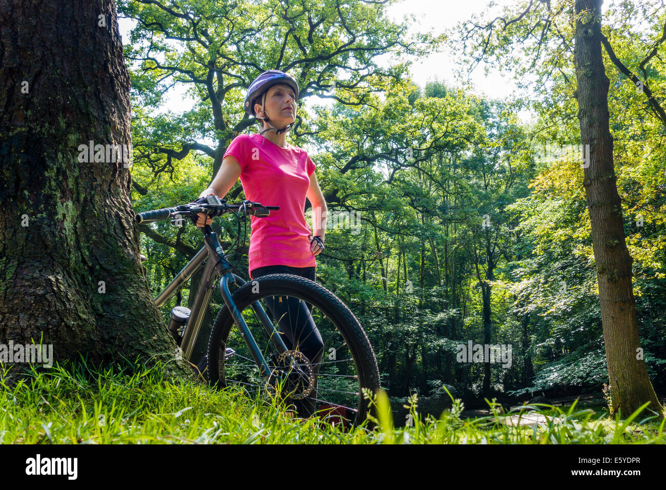 Montare la mezza età della donna nel ciclo di rosa top in piedi con la sua bicicletta nel soleggiato parco alberato di Castell Coch, South Wales UK Foto Stock
