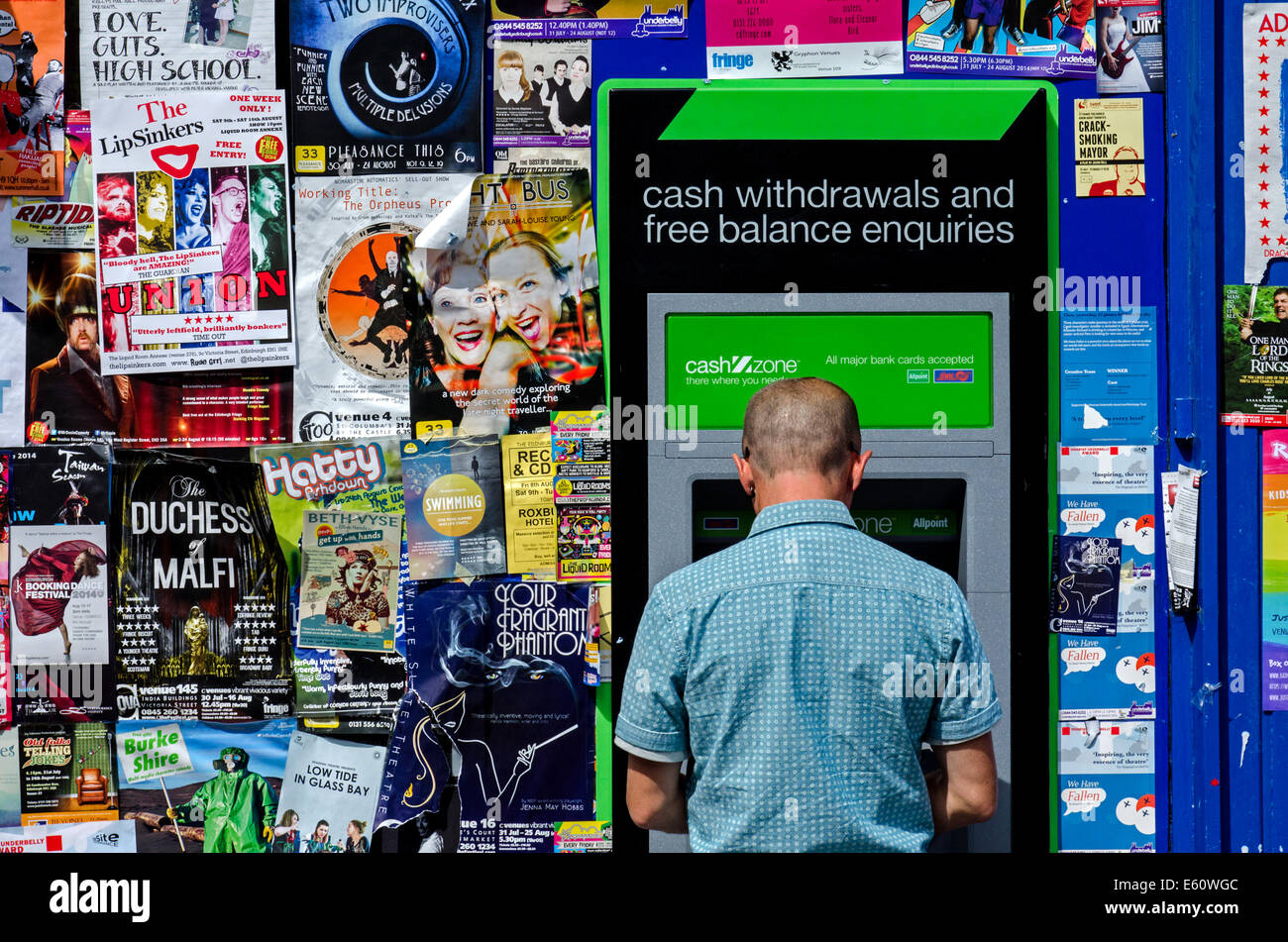 Un giovane uomo con un bancomat in Grassmarket circondato da manifesti con mostra sull'Edinburgh Festival Fringe. Foto Stock