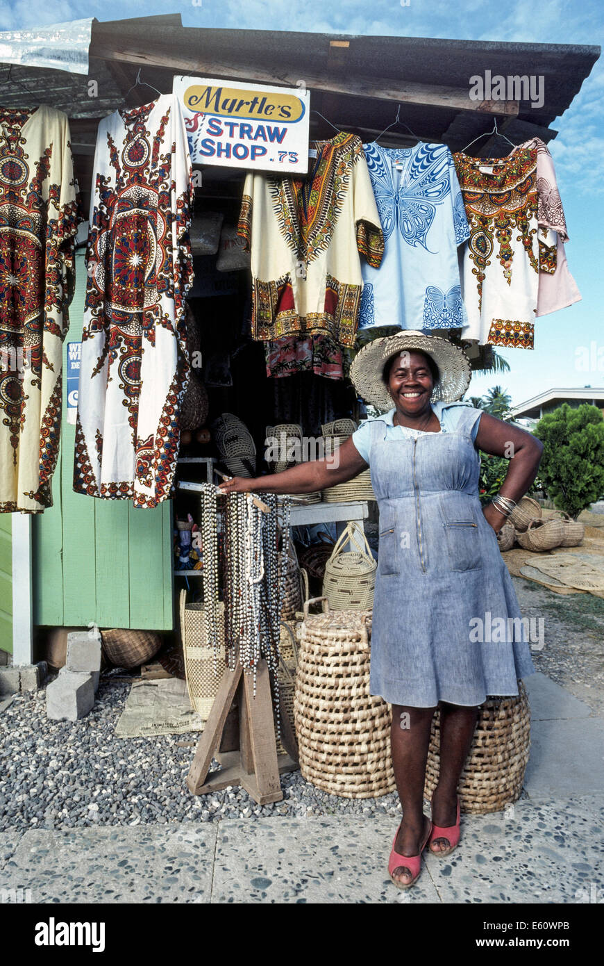 Il Sorridente proprietario di mirto del negozio di paglia mostra il suo ceste, cappelli e altra mercanzia in una Caraibi all'aperto il mercato di artigianato di Ocho Rios, Giamaica. Foto Stock