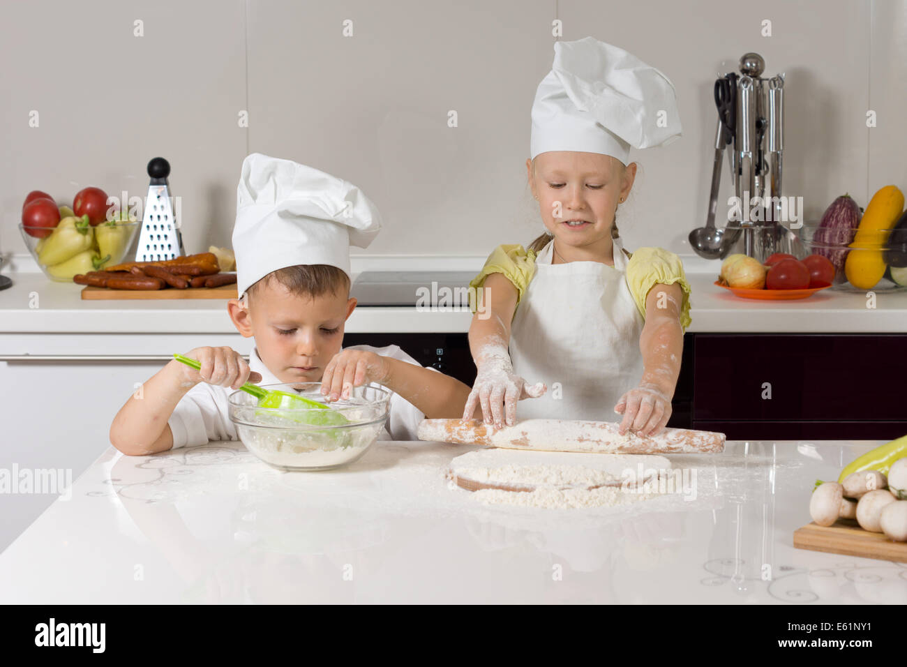 I figli piccoli di cottura di pizze fatte in casa di stendere la pasta per la base in corrispondenza di un banco di cucina nel loro grembiule bianco e gli chef cappelli Foto Stock