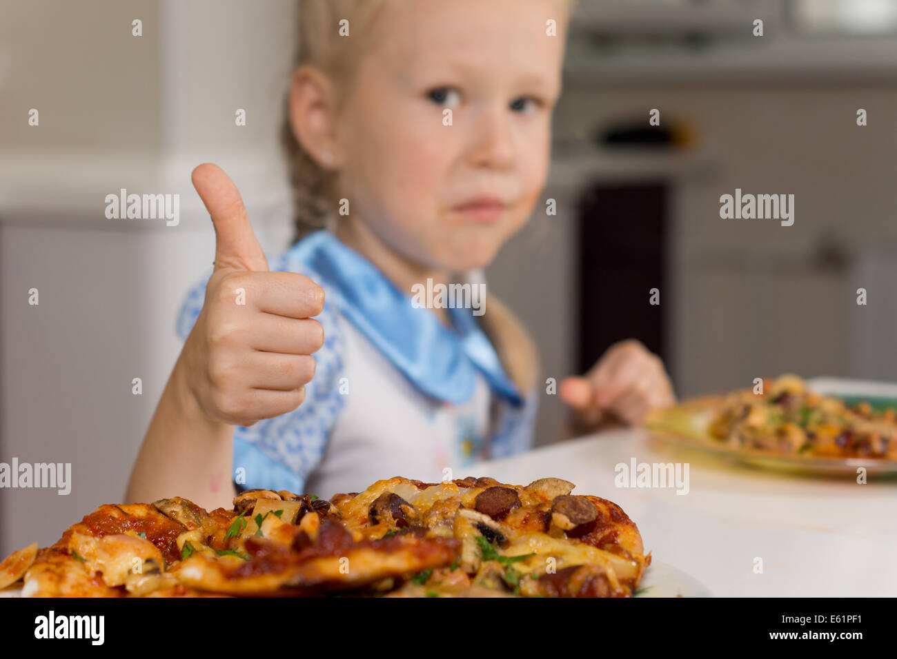 Piccola ragazza godendo di una piastra di pizze dando un pollice in alto di approvazione al segnale la sua approvazione e soddisfazione con un tast Foto Stock
