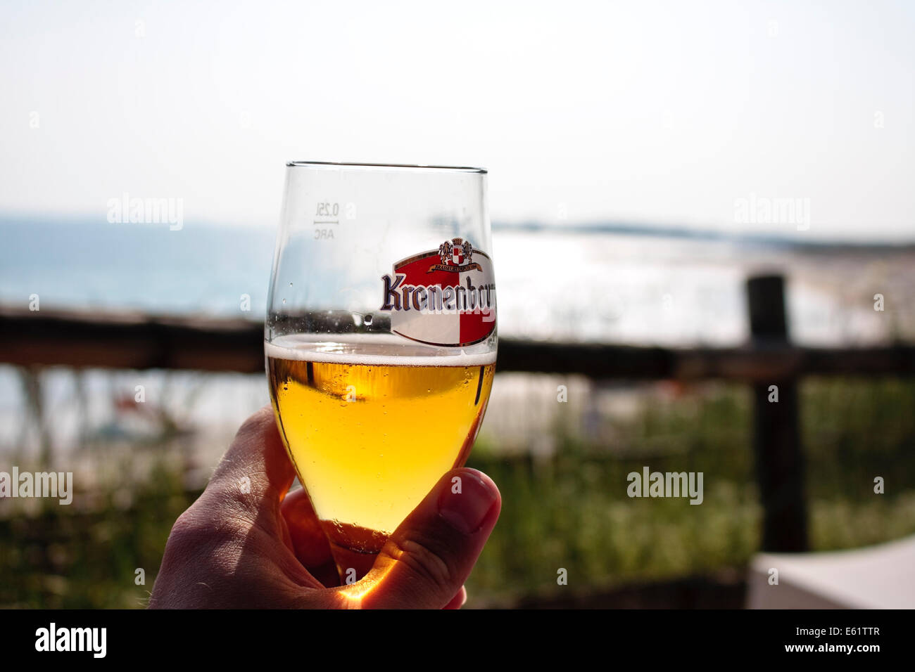 Una mano che tiene un bicchiere di birra Kronenbourg contro una spiaggia soleggiata sfondo Foto Stock