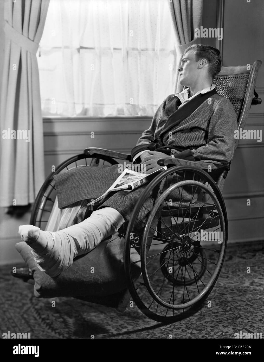 1940s uomo in carrozzella fasciata la gamba e il piede a guardare fuori dalla finestra Foto Stock