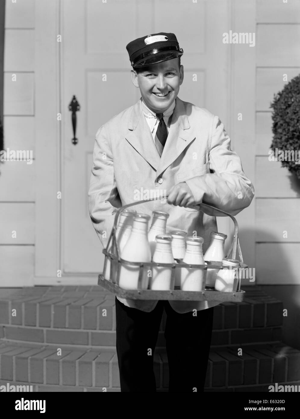 1930s sorridente lattaio guardando la telecamera consegnare latte IN BOTTIGLIE DI VETRO PORTA ANTERIORE Foto Stock