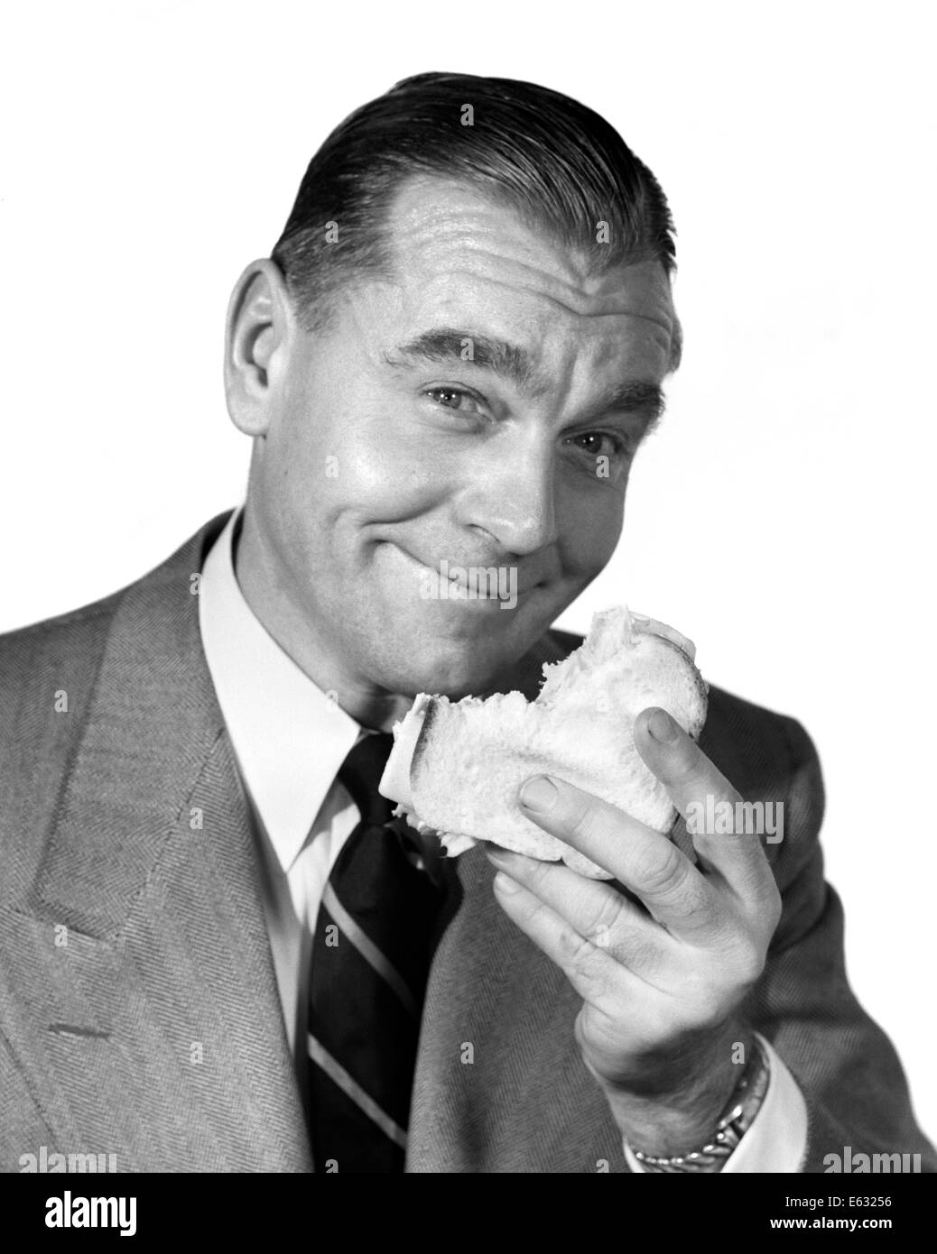 Anni sessanta sorridente imprenditore indossare tuta e cravatta mangiare panino guardando la fotocamera Foto Stock