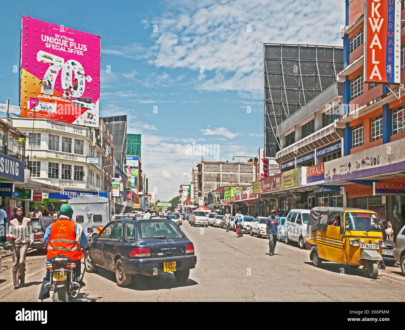 Traffico di persone e di veicoli su Kenyatta Avenue Nakuru Kenya Africa orientale con i pannelli pubblicitari negozi e Piaggio Ape a tre ruote taxi Foto Stock