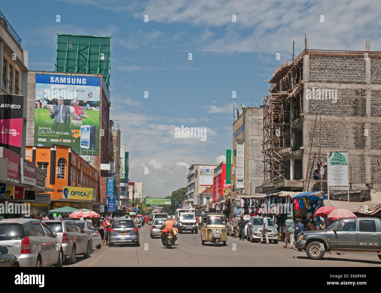 Traffico di persone e di veicoli su Kenyatta Avenue Nakuru Kenya Africa orientale con i pannelli pubblicitari negozi e Piaggio Ape a tre ruote taxi Foto Stock