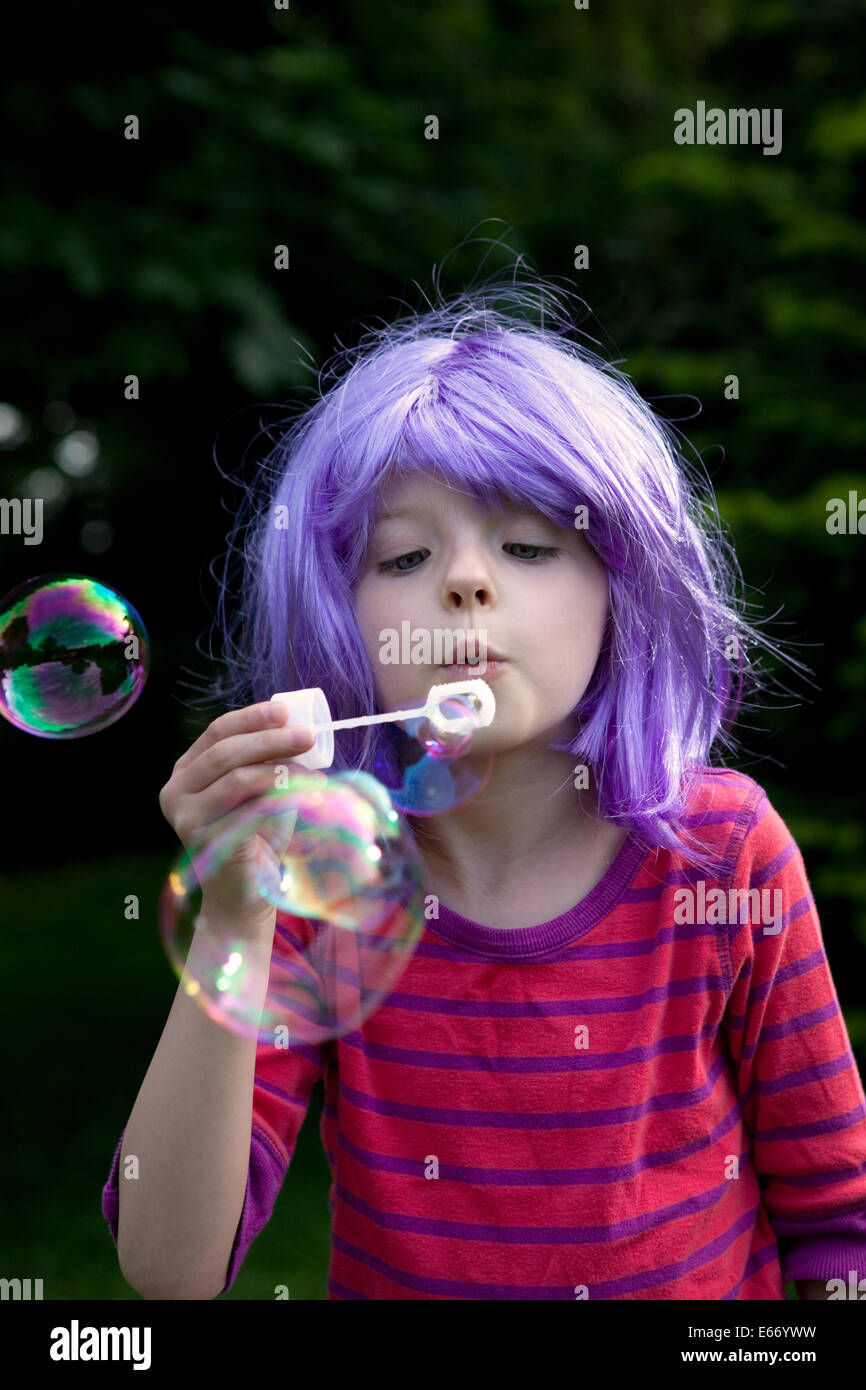 Giovane ragazza che indossa una parrucca viola e soffiare bolle all'esterno. Foto Stock