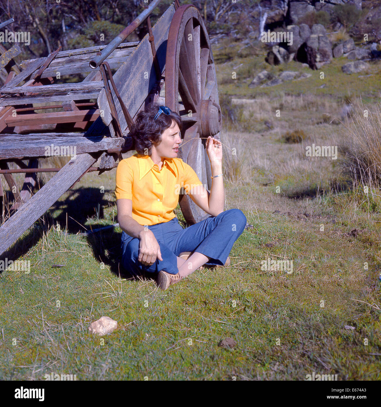 La donna in giallo camicetta e jeans blu masticare la paglia seduti sulla terra da vecchia fattoria carrello Foto Stock