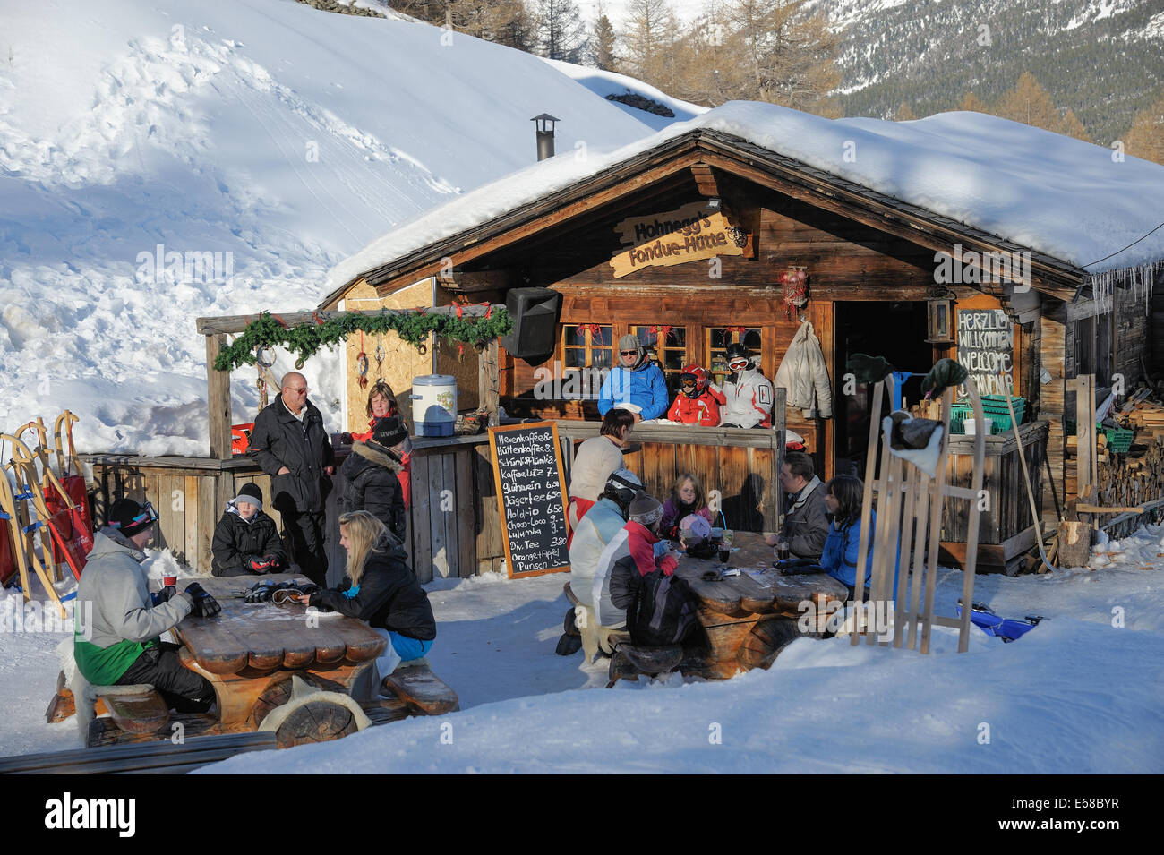 Piccolo ristorante con fonduta svizzera. I clienti stanno avendo un drink all'aperto nella neve approfittando degli ultimi raggi di sole Foto Stock