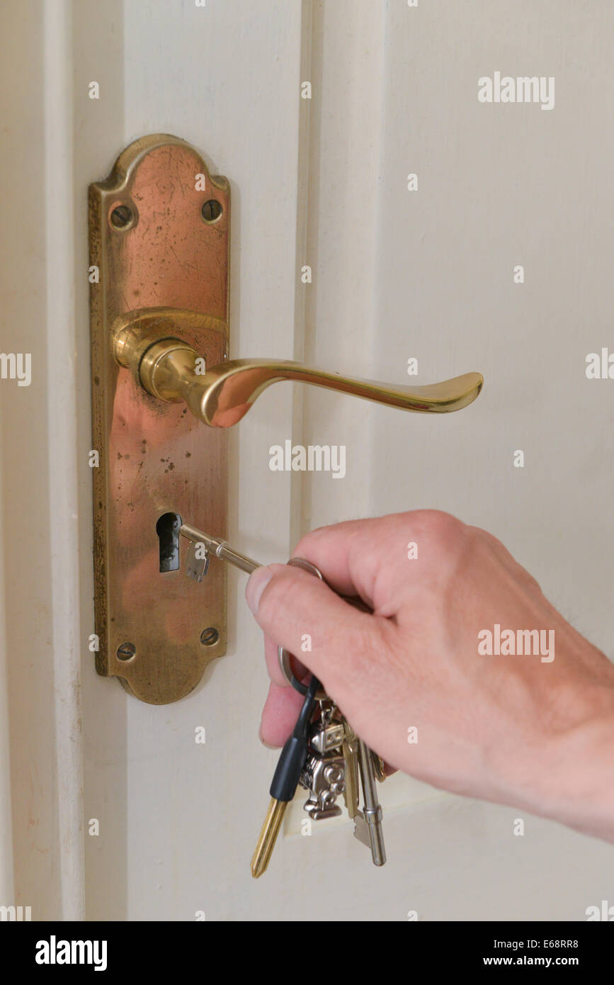 Una mano d'uomo mettendo una chiave in una serratura di una porta bianca all'interno di una casa con una lastra di ottone e maniglia Foto Stock
