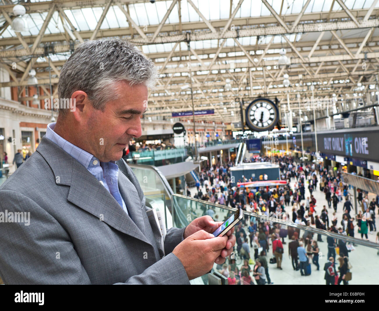 Imprenditore maturo utilizzando un iPhone in Stazione Waterloo di Londra con segnale di occupato atrio della stazione in background Foto Stock