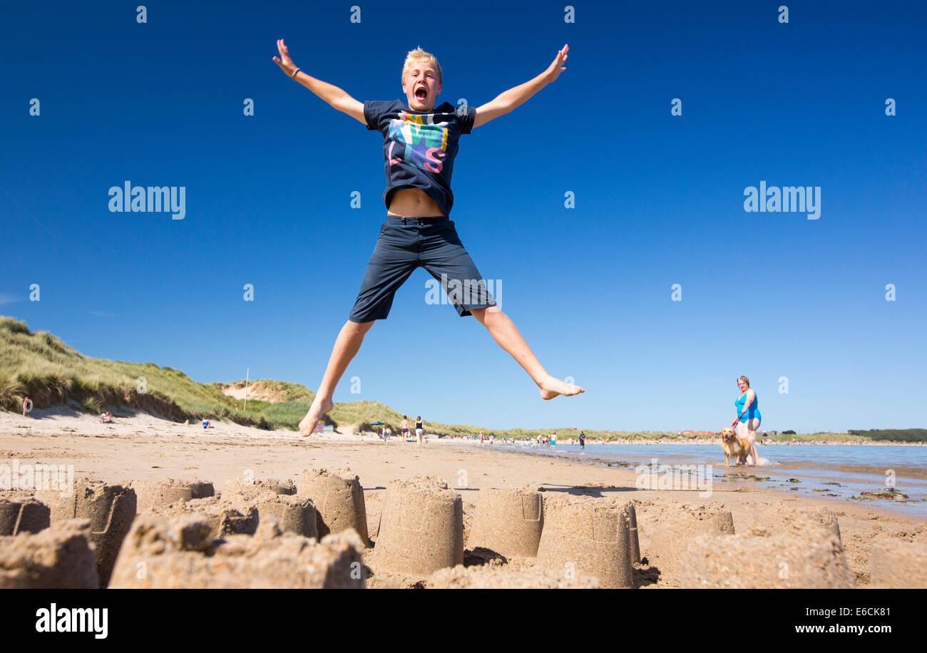 Un ragazzo saltando su un castello di sabbia sulla spiaggia di Bamburgh, Northumberland, Regno Unito. Foto Stock