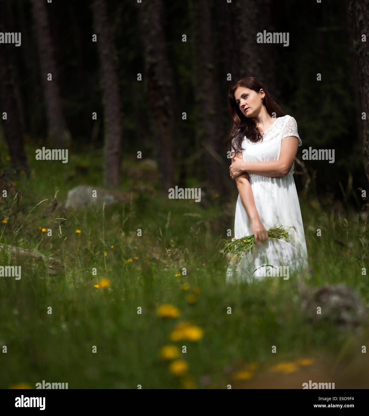 Donna con capelli lunghi in abito bianco e bianco il cappello in mano a piedi nella foresta tenendo un mazzo di fiori Foto Stock