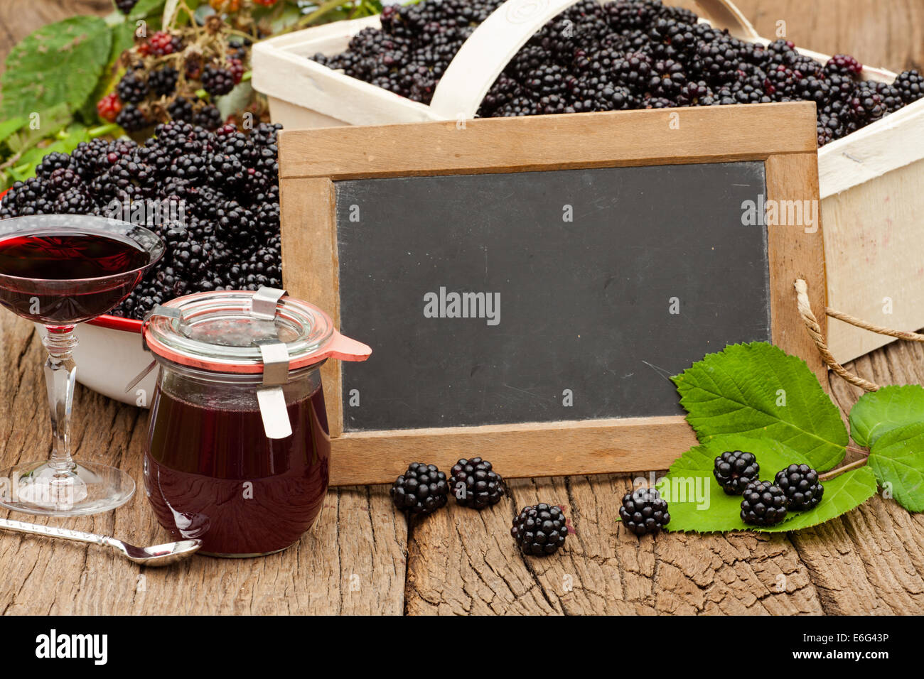 Blank slate blackboard nella parte anteriore del more mature, blackberry liquore e un vasetto di gelatina di blackberry su una tavola in legno rustico Foto Stock